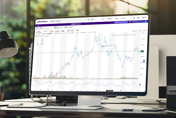 A computer sitting on a desk with a glass window and a lush scene in the background displaying Yahoo Finance charts.