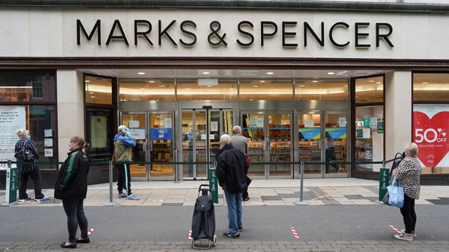 Several shoppers walk along a city street in front of a Marks & Spencer storefront.