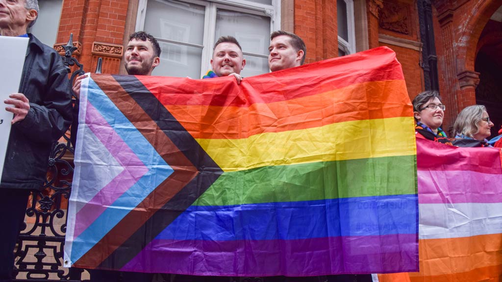 Three people are seen holding up a Progress Pride flag in front of a brick building