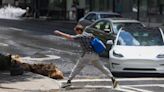 A man leaps to jump over water spilled from a fire hydrant spilled up West Peachtree Street as he crosses 14th Street, Monday, June 3, 2024, in Atlanta.