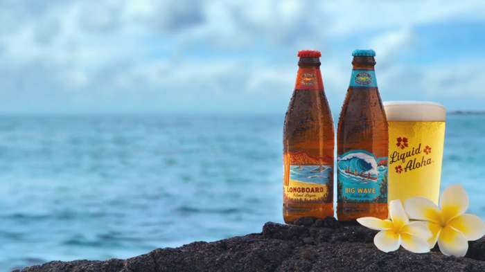 Two bottles and a glass of Kona beer sit on a rock next to a body of water