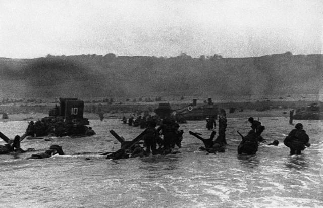 Some of the first assault troops to hit the beachhead hide behind enemy beach obstacles to fire on the Germans, others follow the first tanks plunging through water towards the Normandy shore on June 6, 1944.