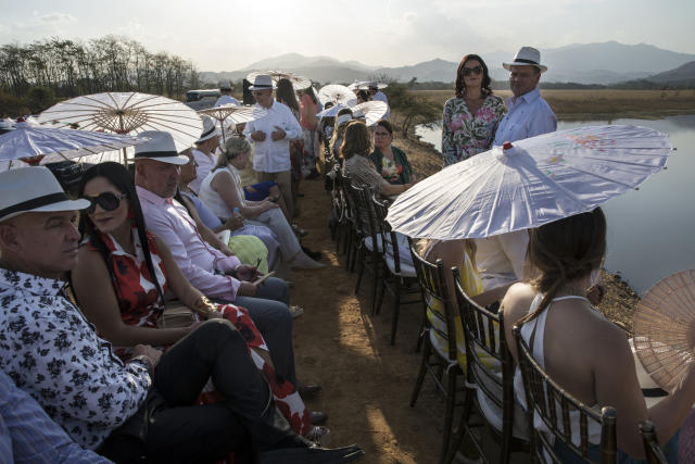 In this Feb. 16, 2019 photo, guests wait for the arrival of groom Juan Jose Pocaterra and his bride Maria Fernanda Vera to their wedding in Acarigua, Venezuela. Women wore long dresses and used colorful umbrellas and folding fans to keep cool under the sun; men followed the recommended attire of white shirts, suspenders and beige pants, and donned Panama hats provided by the organizers. (AP Photo/Rodrigo Abd)