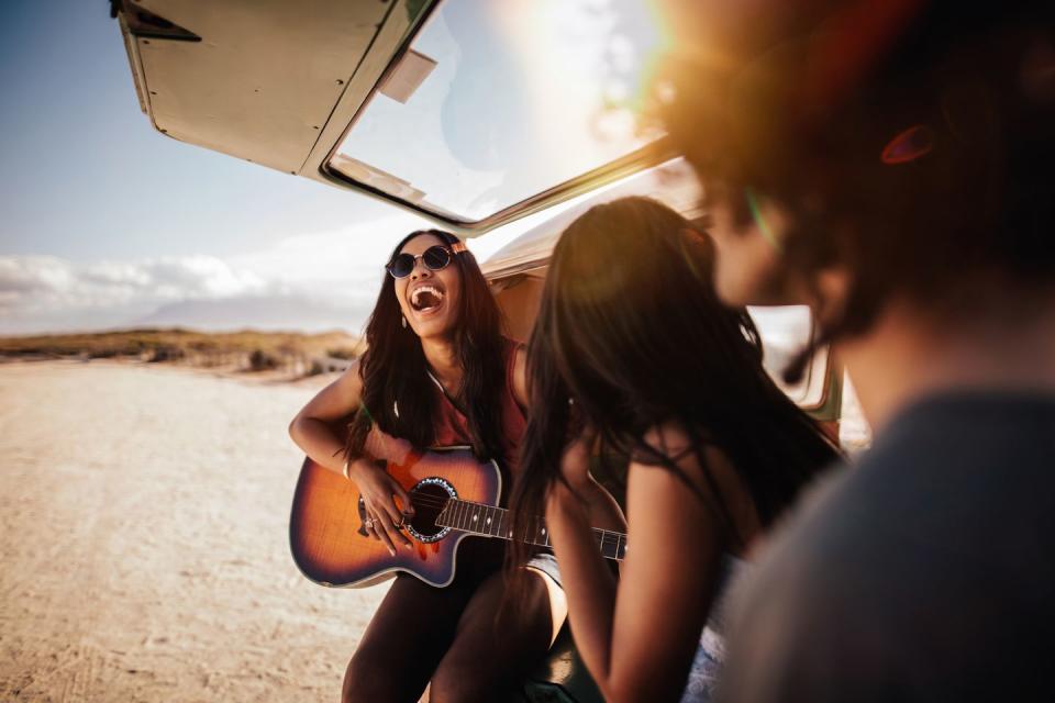 group of friends sit in back of open retro van parked at a sandy beach, playing guitar and smiling at each other on their road trip