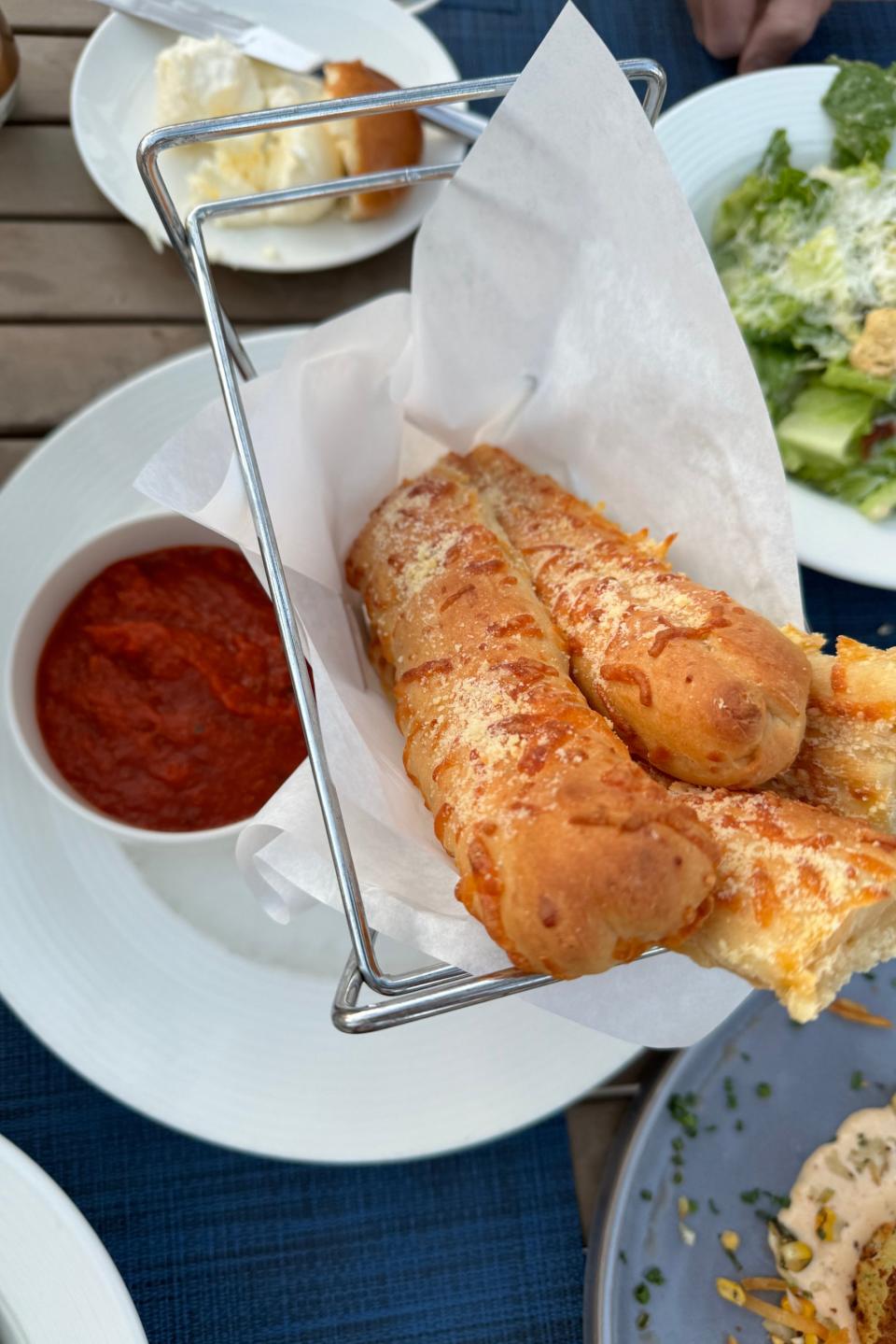 Garlic breadsticks in a metal basket with marinara sauce on a table, suggesting a dining experience while traveling