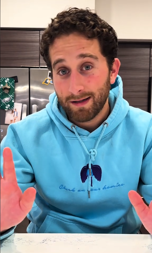 A person in a blue hoodie gestures with open hands in a kitchen setting