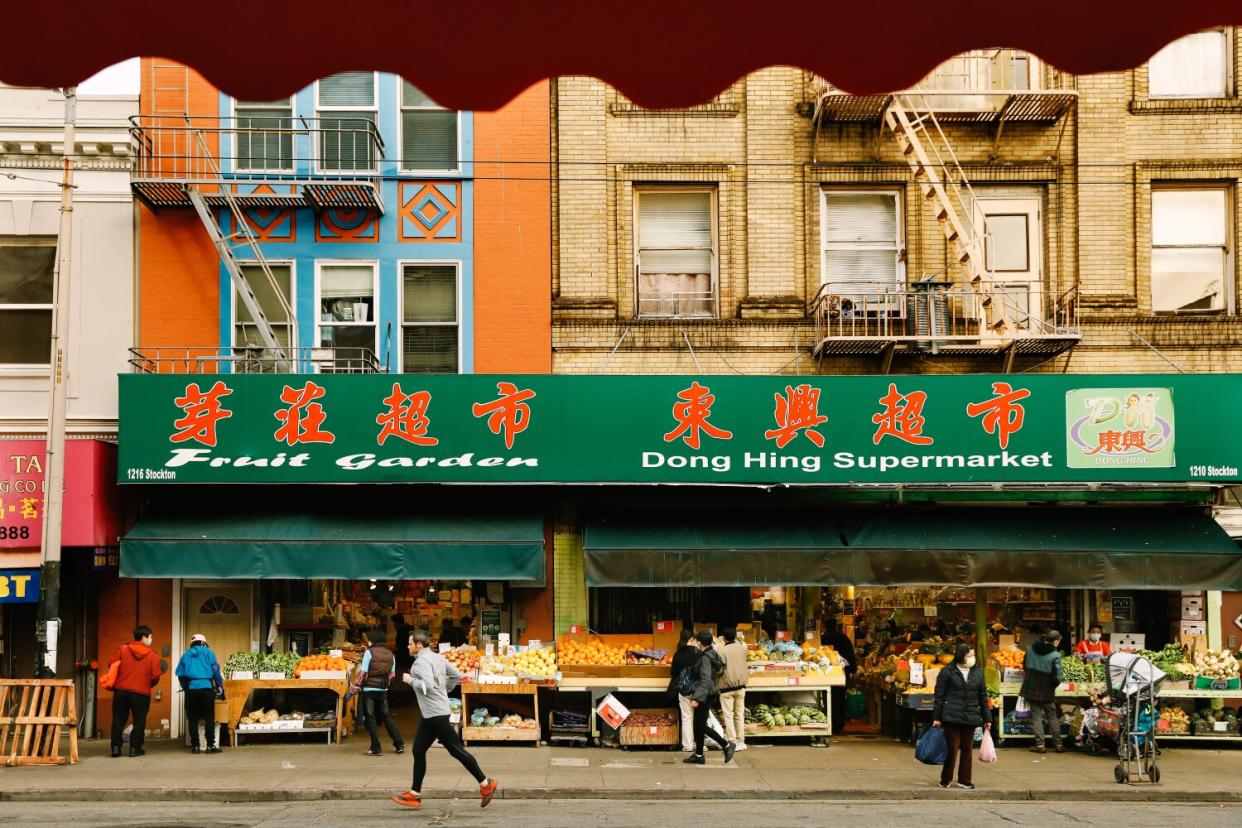 Pedestrians pass a grocery with a sidewalk vegetable stand.