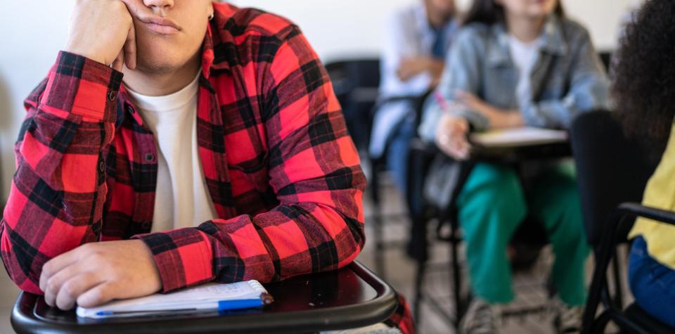 A person wearing glasses and a flannel shirt sits at a school desk with their head resting on their hand, looking bored. Several other students are in the background