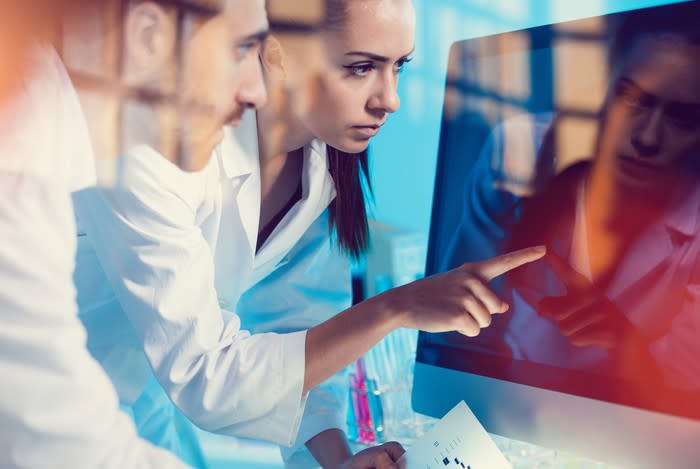 Two people in lab coats looking at a computer monitor.