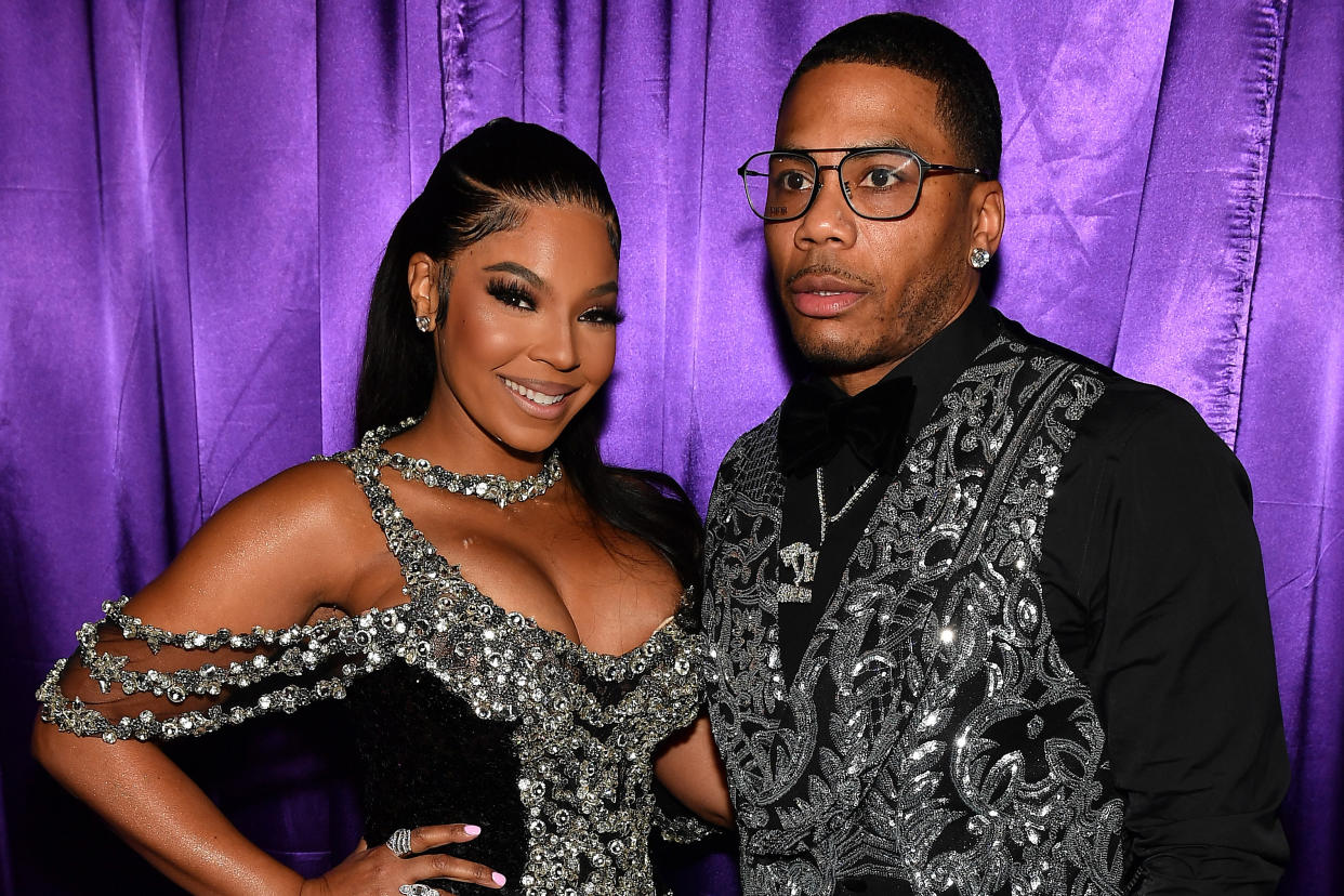 Ashanti and Nelly stand next to each other in front of a purple curtain.