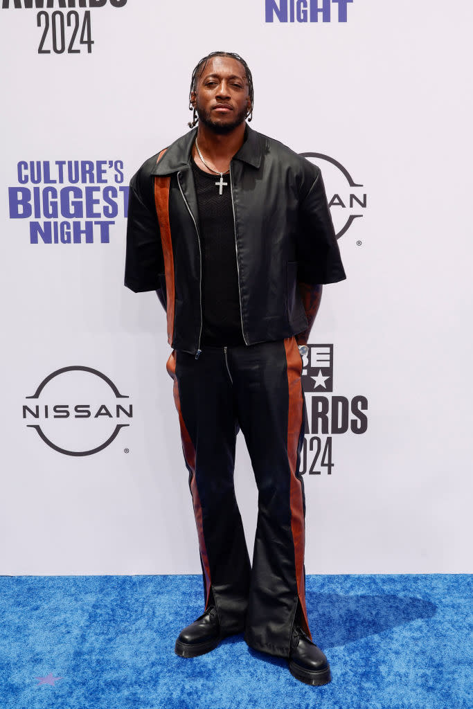 Lecrae, wearing a black jacket and pants with brown stripes, stands on the blue carpet at a BET event