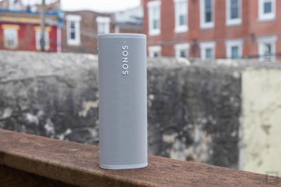 Photo of the Sonos Roam (grayish-white) sitting on a wooden outdoor ledge with old buildings in the distance.