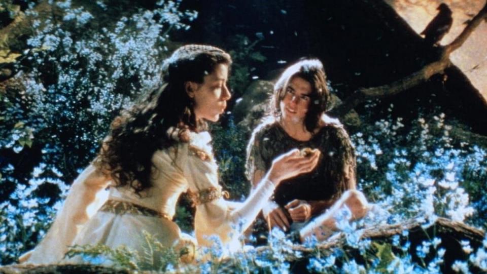 A maiden sits on a meadow of flowers with a long-haired Tom Cruise.