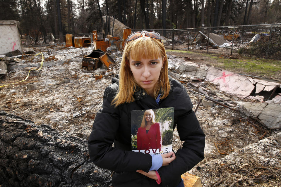 FILE - In this Thursday, Feb. 7, 2019, file photo, Christina Taft poses with a photo of her mother, Victoria Taft, at the burned-out ruins of the Paradise, Calif., home where her mother died the previous fall. Victoria Taft was one of 85 people who died in the most destructive wildfire in California history. Christina, who now lives in San Jose, said it's been a difficult year and is planning to return to Paradise, Friday, Nov. 8, 2019, for the one-year anniversary of the wildfire that took her mother's life. (AP Photo/Rich Pedroncelli, File)