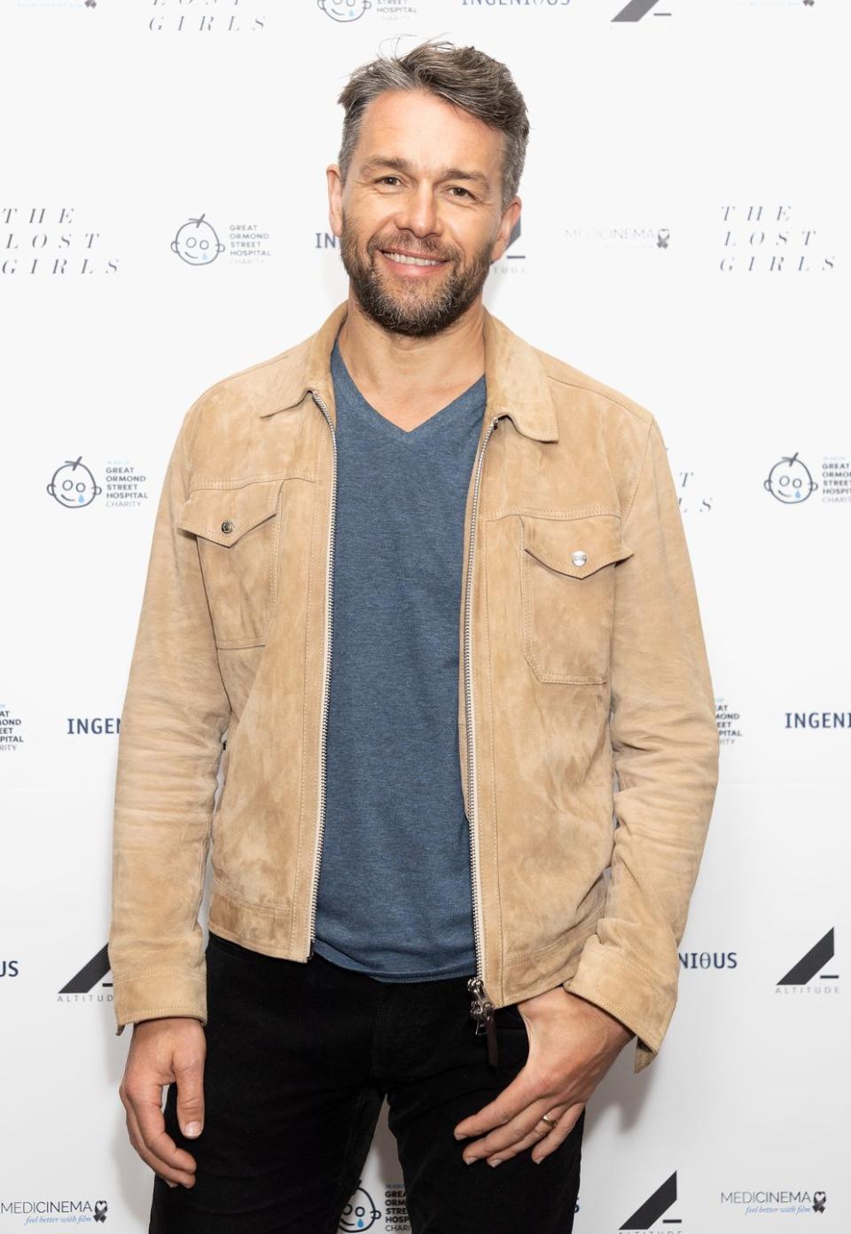 julian ovenden, a man stands smiling at the camera, he has greying brown hair and a short beard, he wears a blue jumper with suede brown jacket and black jeans