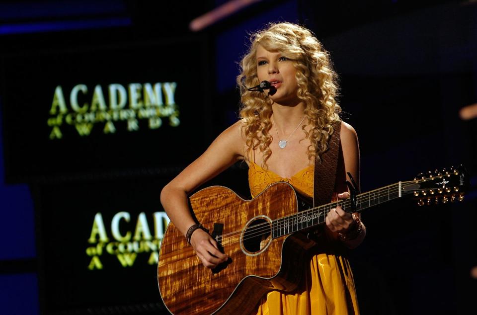 taylor swift sings into a microphone headset as she plays acoustic guitar on a stage, she wears a yellow sleeveless dress and silver necklace