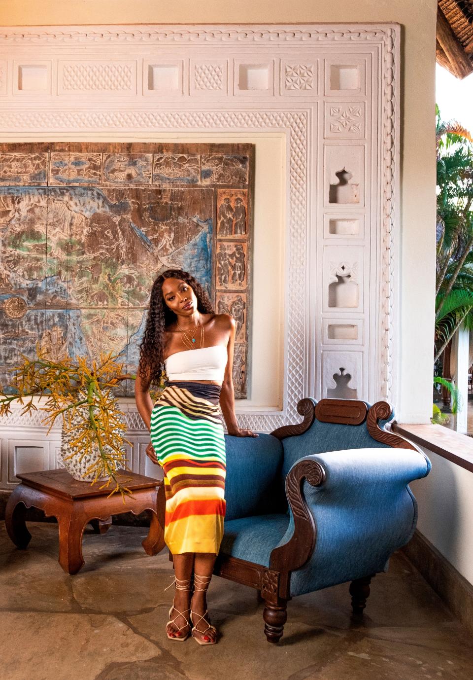 Naomi Campbell, wearing a Jade Swim bandeau top, Dries van Noten skirt, and Cult Gaia sandals, in the entrance area of her Kenyan getaway. On wall, a Bas-relief of Africa by local artist Armando Tanzini. Fashion styling by Carlos Nazario.