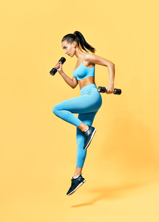 Sporty woman in sportswear jumping with dumbbells on yellow background. Strength and motivation concept. Full length profile portrait