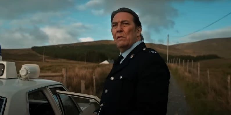 ciaran hinds in the land of saints and sinners movie, his character wears a police uniform and looks thoughtful as he stands in front of some fields