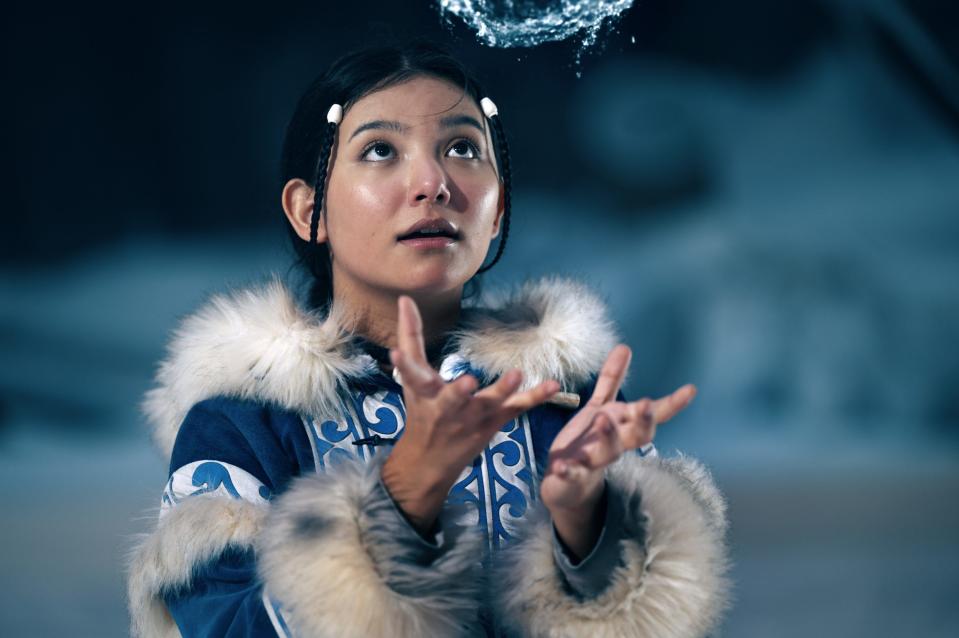 katara in the live-action netflix avatar. she's wearing blue, patterned robes with fur on the cuffs and collar, her hair pulled back with two strands looping in the front. her hands are outstretched, and she's looking at a ball of water she's making hover in the air