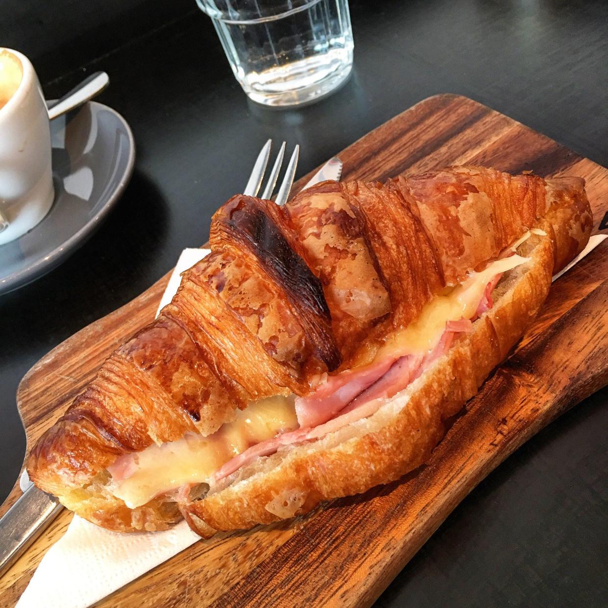 Ham and cheese croissant served on a wooden board with an espresso and a small glass of water.