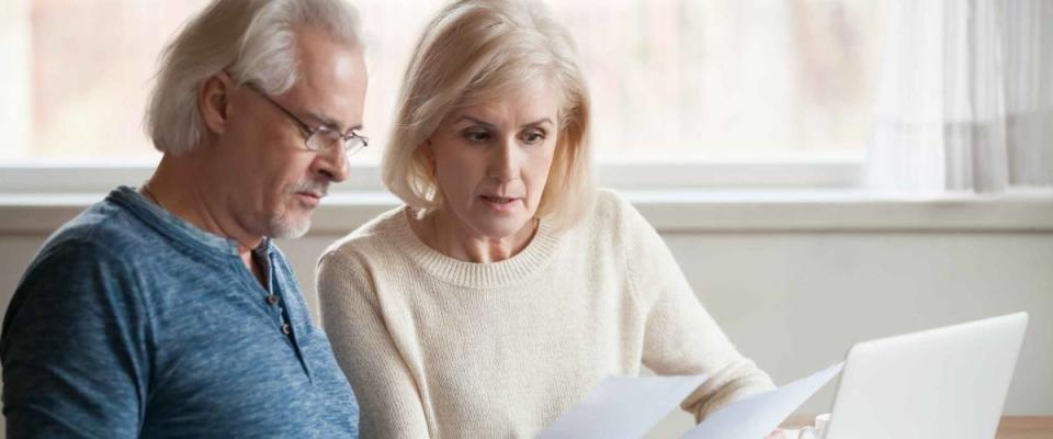 Serious looking older couple look at pieces of paper at kitchen table.