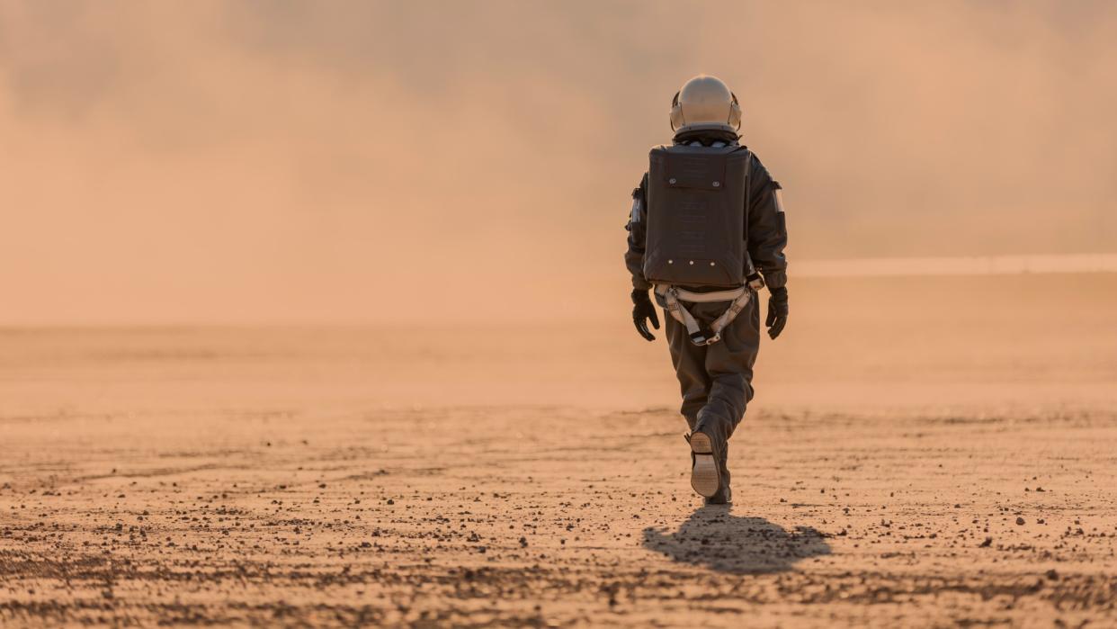  A person in a brown spacesuit walking on a reddish-orange desert. 