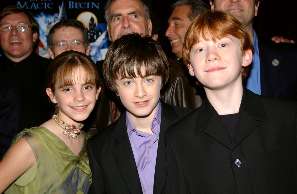 Emma Watson, Daniel Radcliffe, and Rupert Grint at a public event, dressed in semi-formal attire, posing for a photo with other people in the background