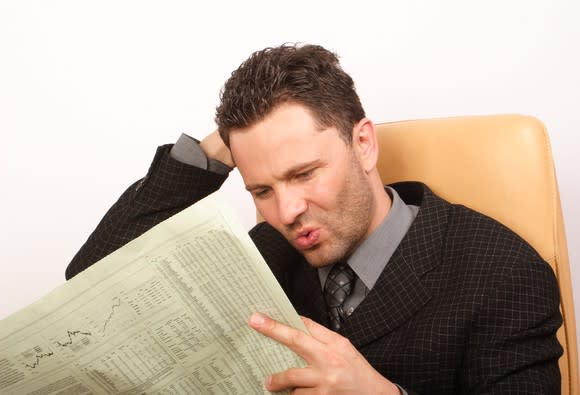 A seated man in a suit reading a financial newspaper, searching for undervalued stocks with his mouth puckered.