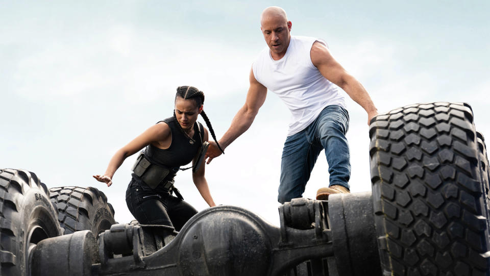 Vin Diesel and Nathalie Emmanuel stand on top of a large overturned vehicle.