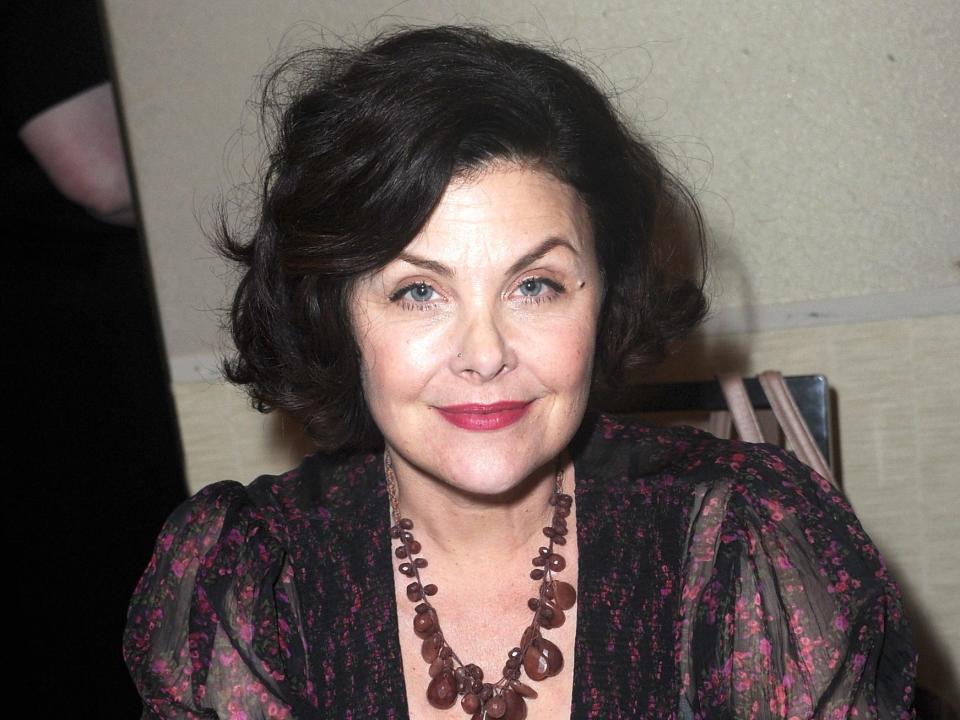 sherilyn fenn sitting at a table with her arms resting on the top, looking towards the camera and smiling