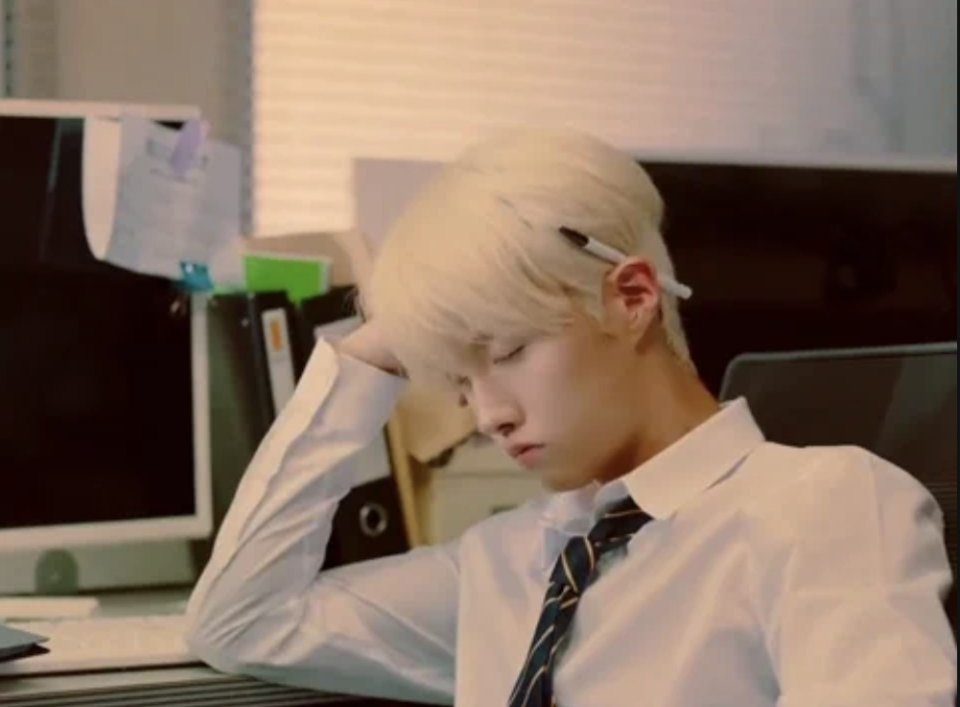 A person with blonde hair wearing a white shirt and tie is asleep at a desk in an office, with a pen in their hair