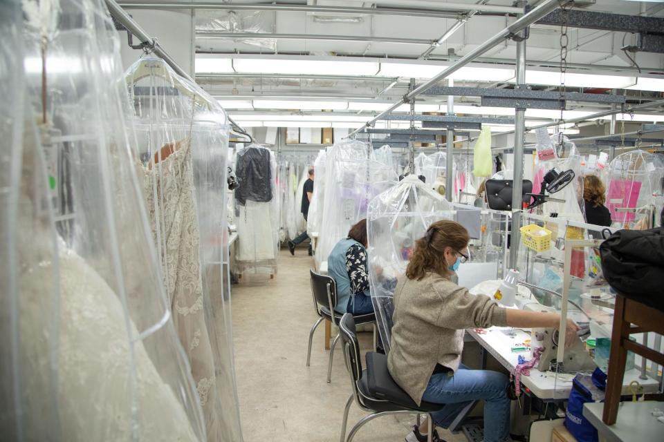 Women sew gowns at desks in a room full of wedding dresses.