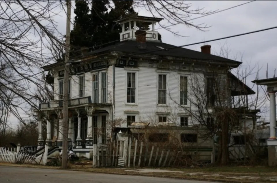 A dilapidated two-story house with a cupola and a collapsed porch surrounded by overgrown vegetation