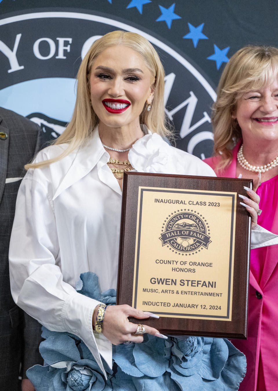 Gwen Stefani holding a plaque at the Orange County Music, Arts & Entertainment Hall of Fame 2023 event, smiling, with two individuals partly visible behind her