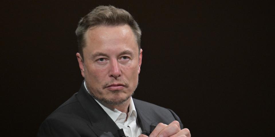 Elon Musk clasping his hands together with a serious expression on his face while wearing a black suit with a white button-down shirt.