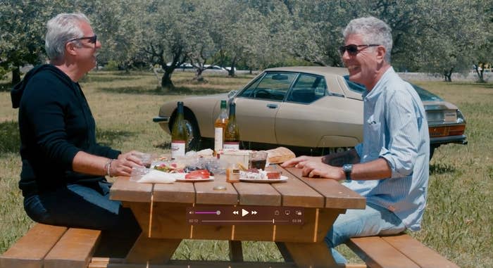Ripert and Bourdain enjoying a picnic lunch outdoors, sitting on a bench