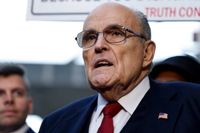 Rudy Giuliani, wearing glasses and a dark suit with a red tie, speaks with people in background