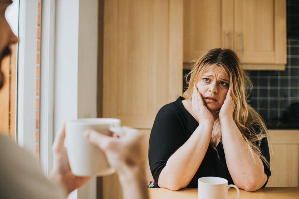 A woman looks anxious as she sits at a kitchen table with her head in her hands