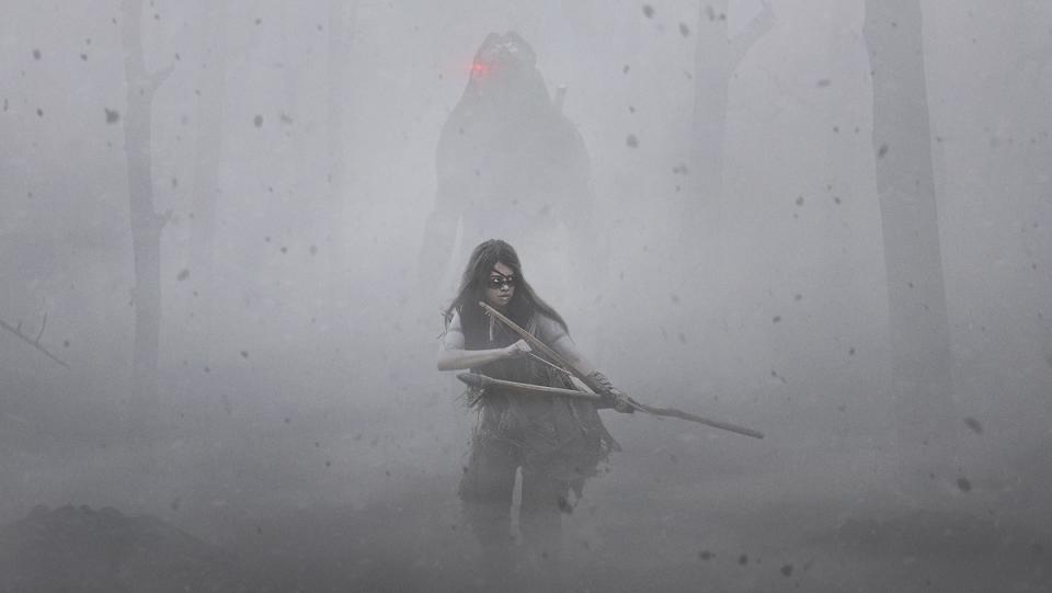 A hazy, misty forest with an Indigenous warrior with a bow and arrow and behind them is the silhouette of a Predator with a glowing red eye piece in the upcoming Predator prequel Prey.