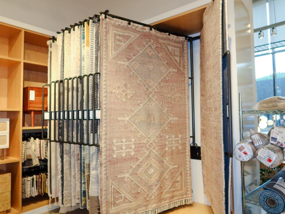 A rug display at West Elm. Several rugs hang from a rack. One rug has a light-red and white geometric design.