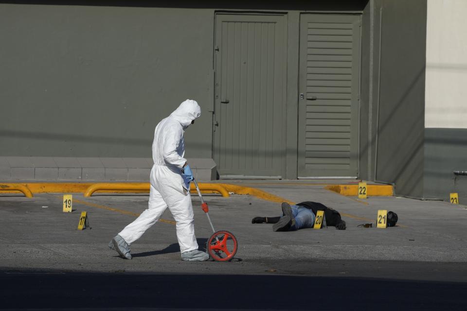 FILE - An attacker lies dead on his back in the parking lot of a convenience store as a police investigator uses a measuring wheel while collecting evidence from a crime scene where a municipal policeman was shot dead, in Celaya, Mexico, Feb. 28, 2024. The policeman had been driving his wife to work when cartel gunmen opened fire on their car. The policeman killed one attacker before dying. (AP Photo/Fernando Llano, File)