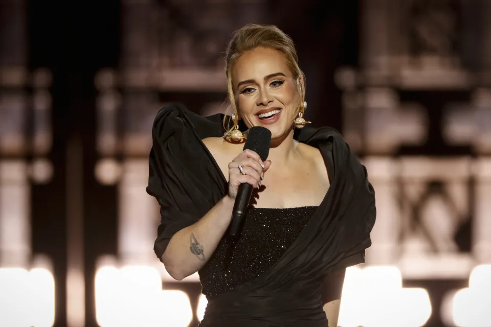 Adele performs with a microphone, wearing a black off-the-shoulder dress with puffed sleeves and large gold earrings