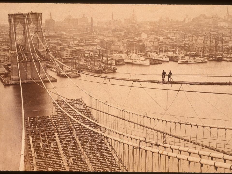 Two men standing on a high catwalk, surveying the construction of the Brooklyn Bridge