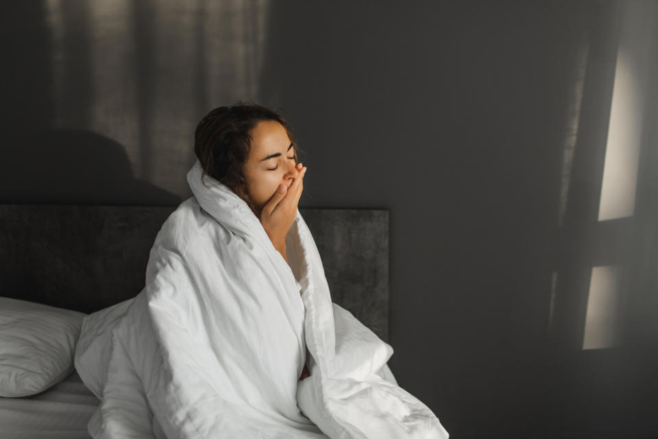 A woman sits in bed wrapped in a blanket, yawning with a hand covering her mouth, in a dimly lit room