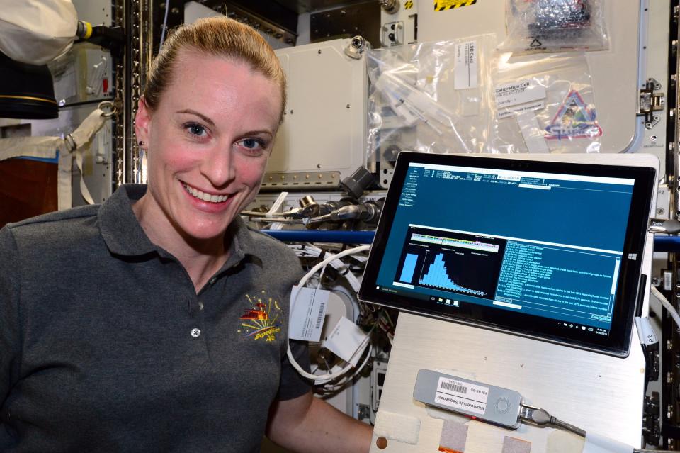 Kate Rubins smiles at the camera while next to a screen showing data and graphs.