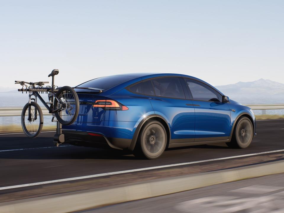 A blue Tesla Model X carrying a bicycle and driving on a road against a blue sky.