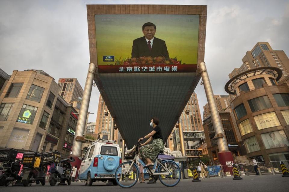 A woman wearing a face mask rides a bicycle past a large television screen at a shopping center displaying Chinese state television news coverage of Chinese President Xi Jinping