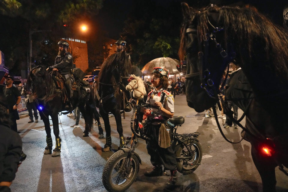 Performer clown Hashoteret Az-Oolay stands next to Israeli police officers on horses as she takes part in a protest against Israeli Prime Minister Benjamin Netanyahu's government, demanding new elections and the release of the hostages held in the Gaza Strip by the Hamas militant group, in Jerusalem, Monday, June 17, 2024. (AP Photo/Ohad Zwigenberg)