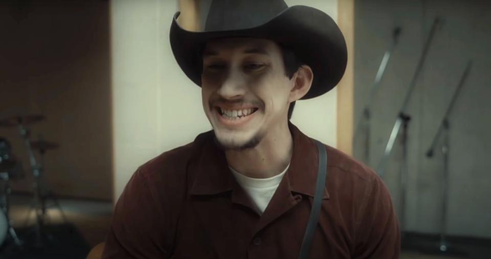 Adam person wearing a cowboy hat and suspenders, sitting indoors and singing